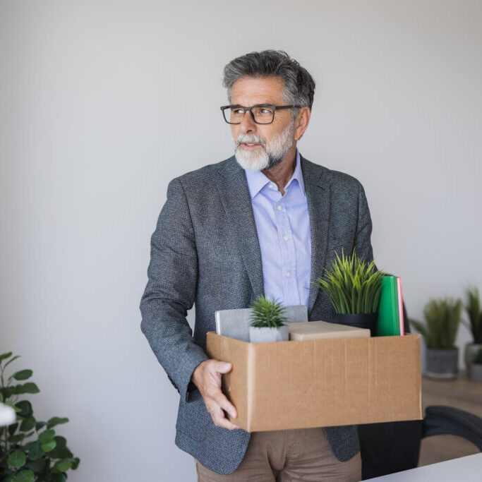 Sad fired mature manager leaving office holding a box with his belongings
